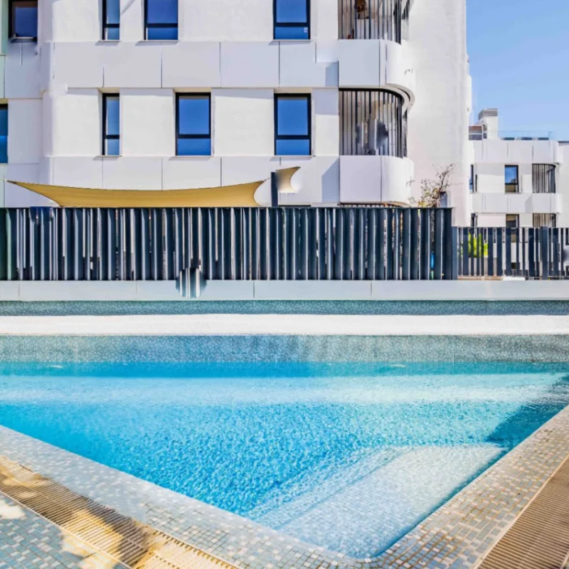 Piscina comunitaria con revestimiento de gresite azul y escaleras integradas, rodeada de una valla de seguridad moderna frente a un edificio de arquitectura contemporánea en Jávea.