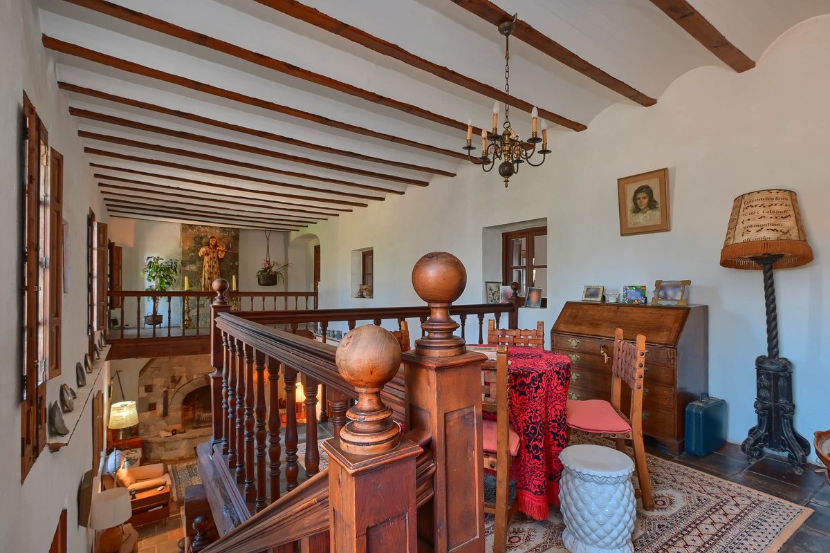 Altillo superior con vigas de madera oscura a la vista, barandilla de madera torneada y zona de despacho con muebles antiguos y vistas al salón inferior con chimenea de piedra.
