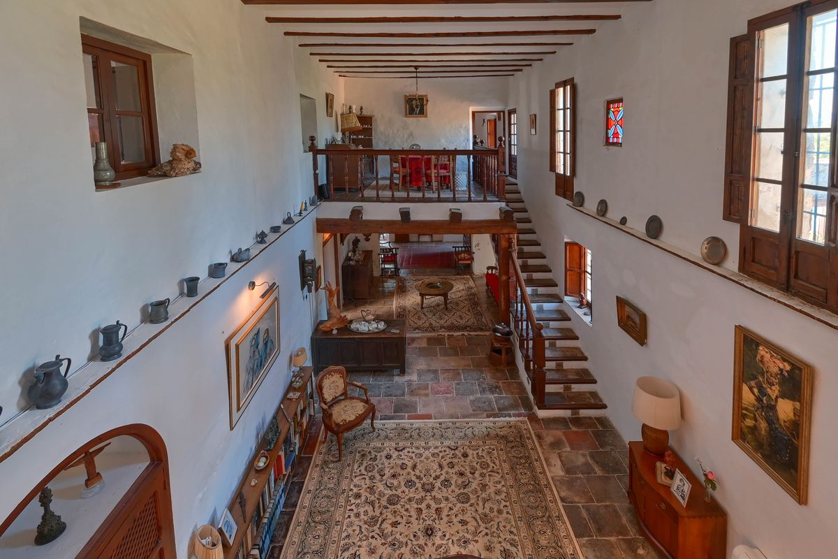 Amplio vestíbulo de doble altura con suelos de barro cocido, vigas de madera a la vista y una escalera que conduce a una galería superior con balaustrada rústica.