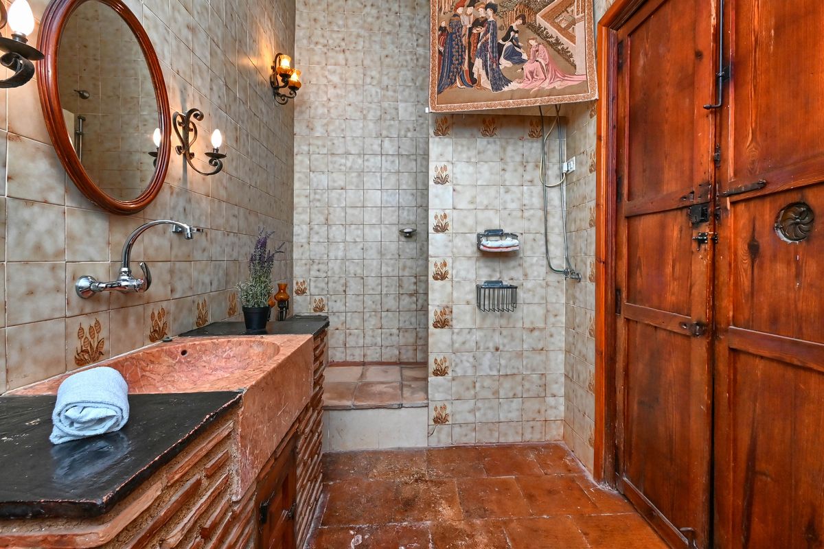 Baño de estilo rústico con lavabo de piedra rosada, paredes de azulejos decorados, apliques de hierro forjado y una puerta de madera maciza tradicional.
