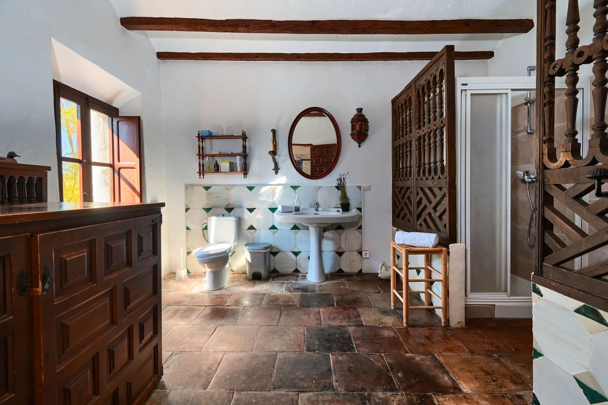Baño rústico con suelos de barro cocido, vigas de madera a la vista y una celosía tallada. Dispone de lavabo de pedestal sobre un frontal de azulejos decorativos.