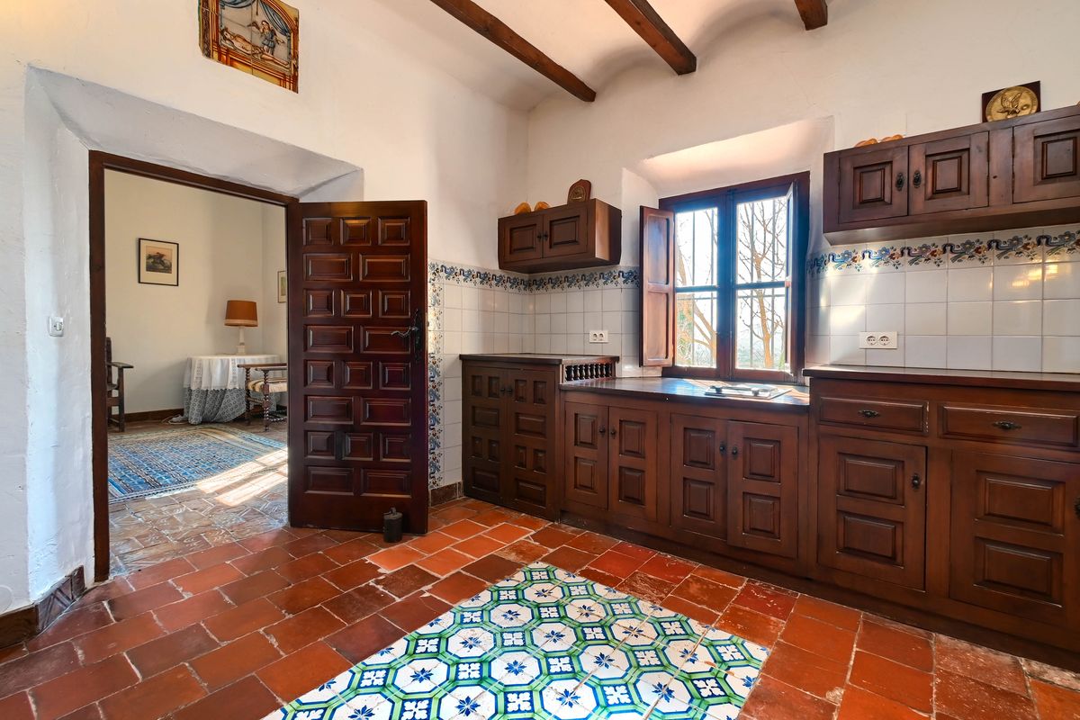 Cocina tradicional con armarios de madera oscura, suelos de terracota con mosaicos decorativos y vigas vistas. Una puerta robusta conecta con la estancia contigua.