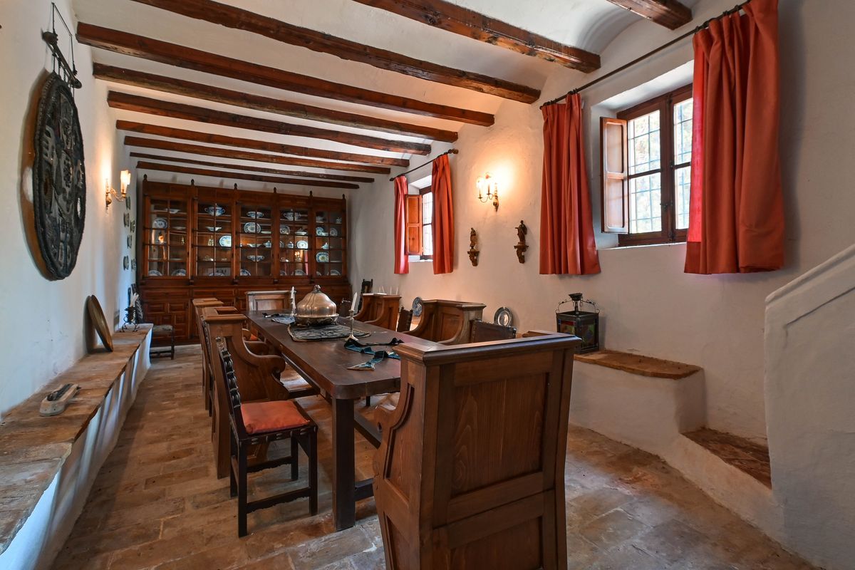 Comedor histórico con vigas de madera vista, suelo de barro cocido y una gran alacena de madera integrada con vajilla tradicional y cerámica.