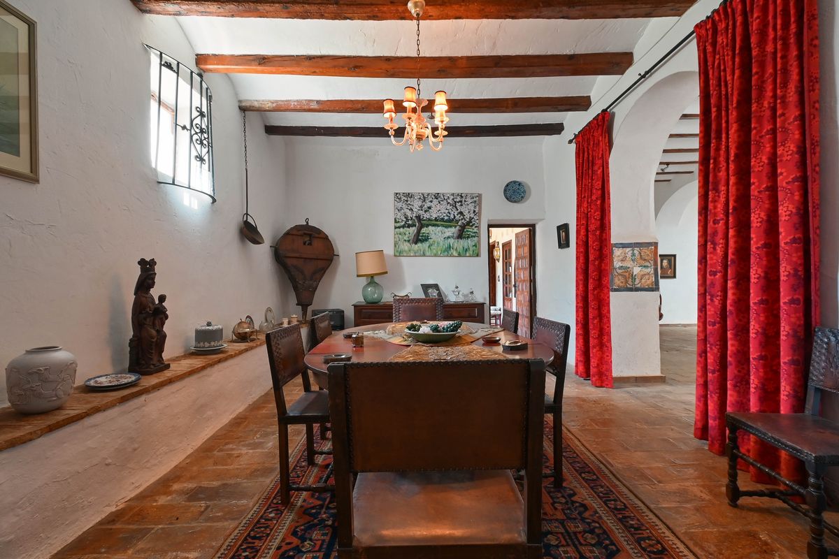 Comedor rústico con vigas de madera a la vista, suelos de terracota y mobiliario de cuero tradicional junto a un arco con cortinas rojas.