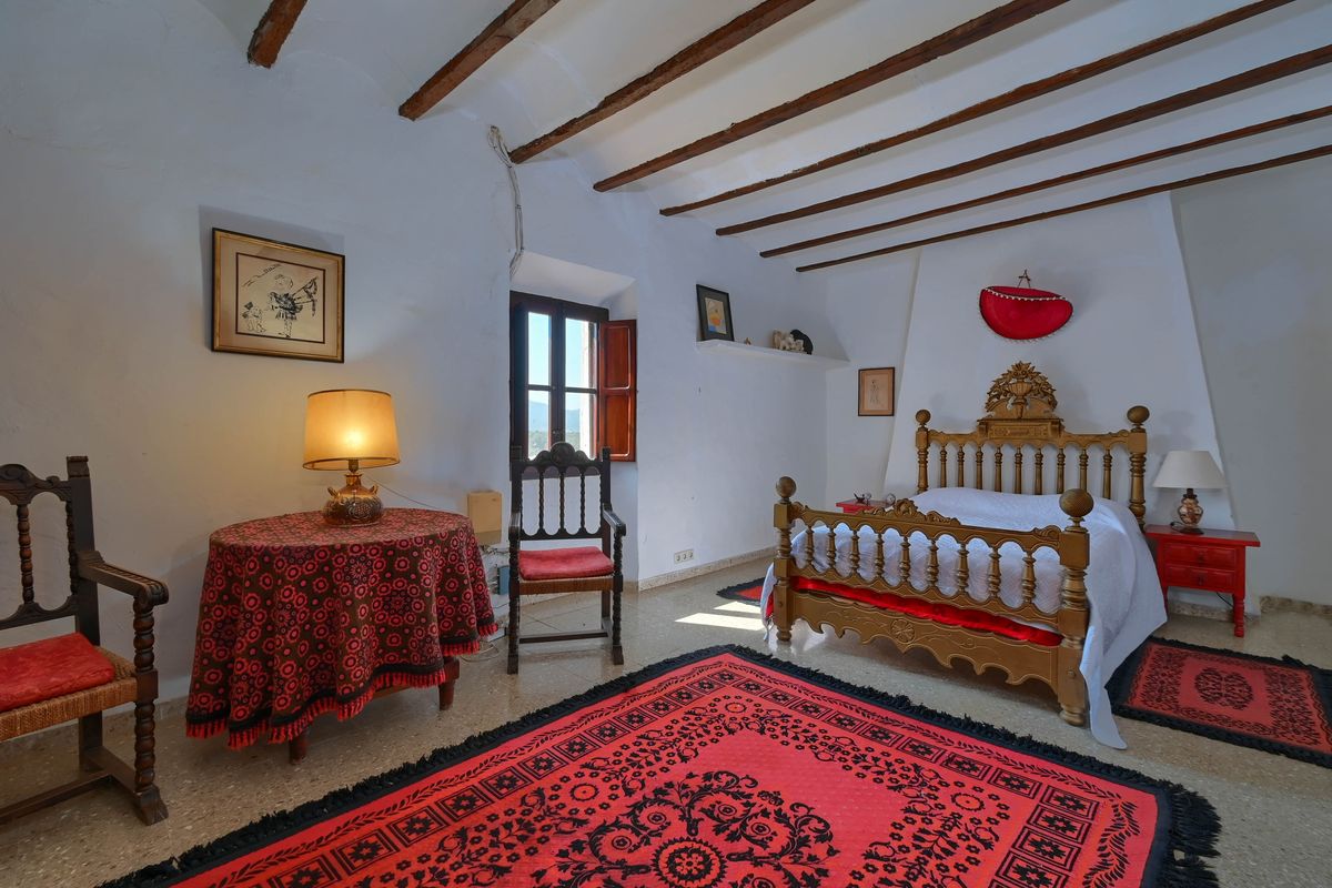 Dormitorio de estilo rústico con vigas de madera originales, cama tallada artesanalmente y alfombras rojas decorativas sobre suelo de piedra.