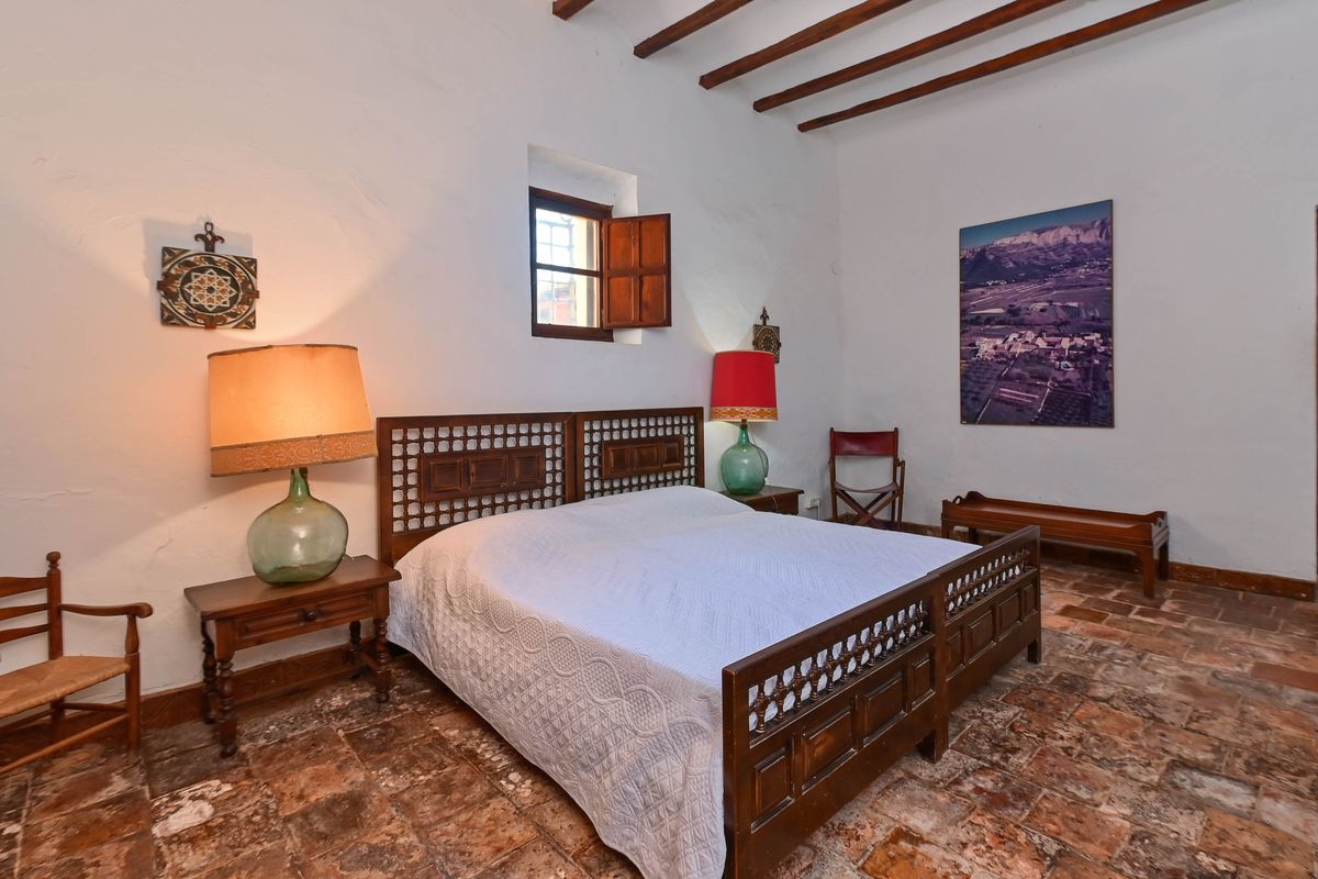 Dormitorio rústico con suelos de barro cocido, vigas de madera vista y cama con cabecero tallado. Incluye lámparas de damajuana y ventana tradicional con contraventanas de madera.