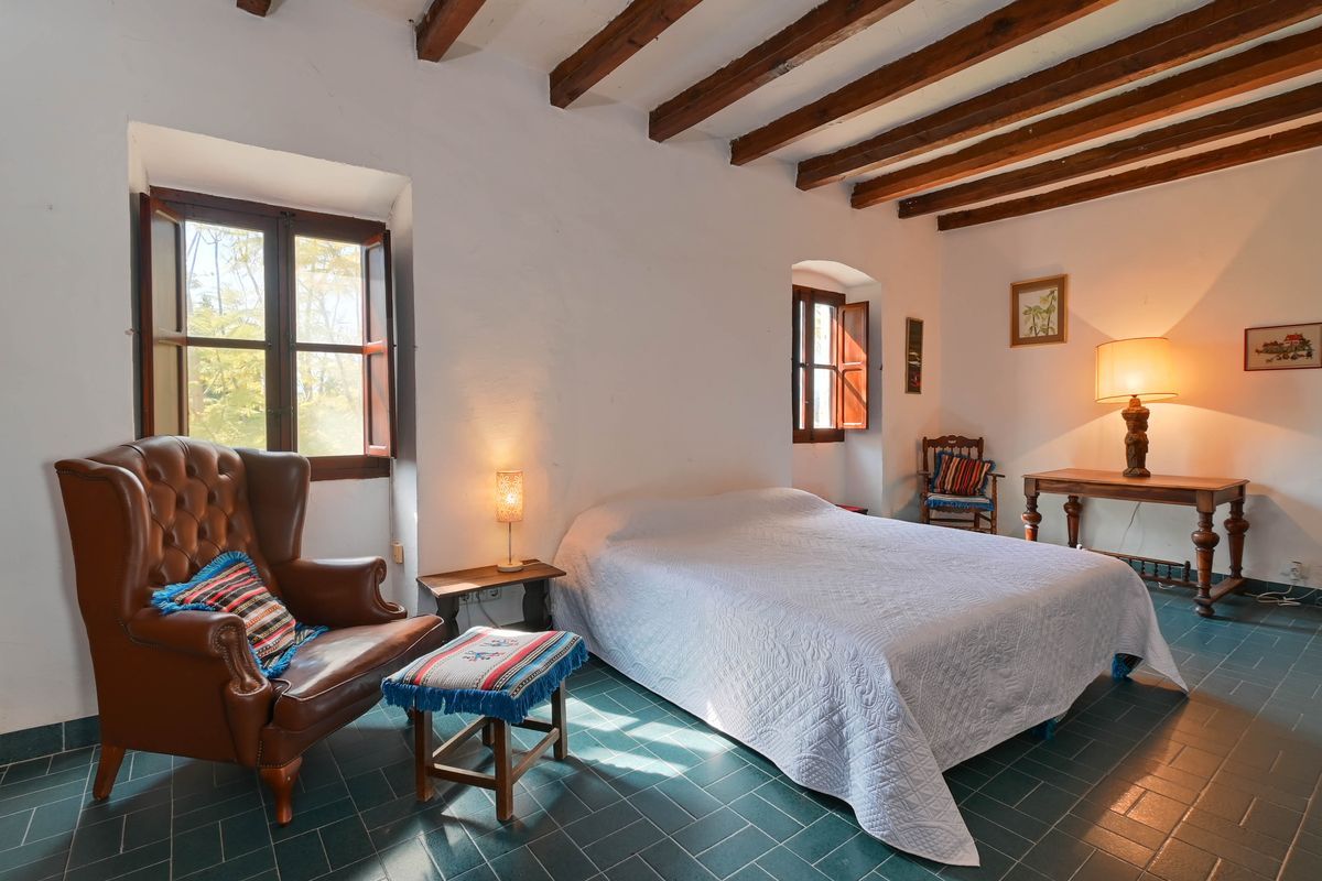 Dormitorio rústico con vigas de madera a la vista, suelos de baldosa verde y un sillón orejero de cuero junto a ventanas tradicionales con contraventanas.