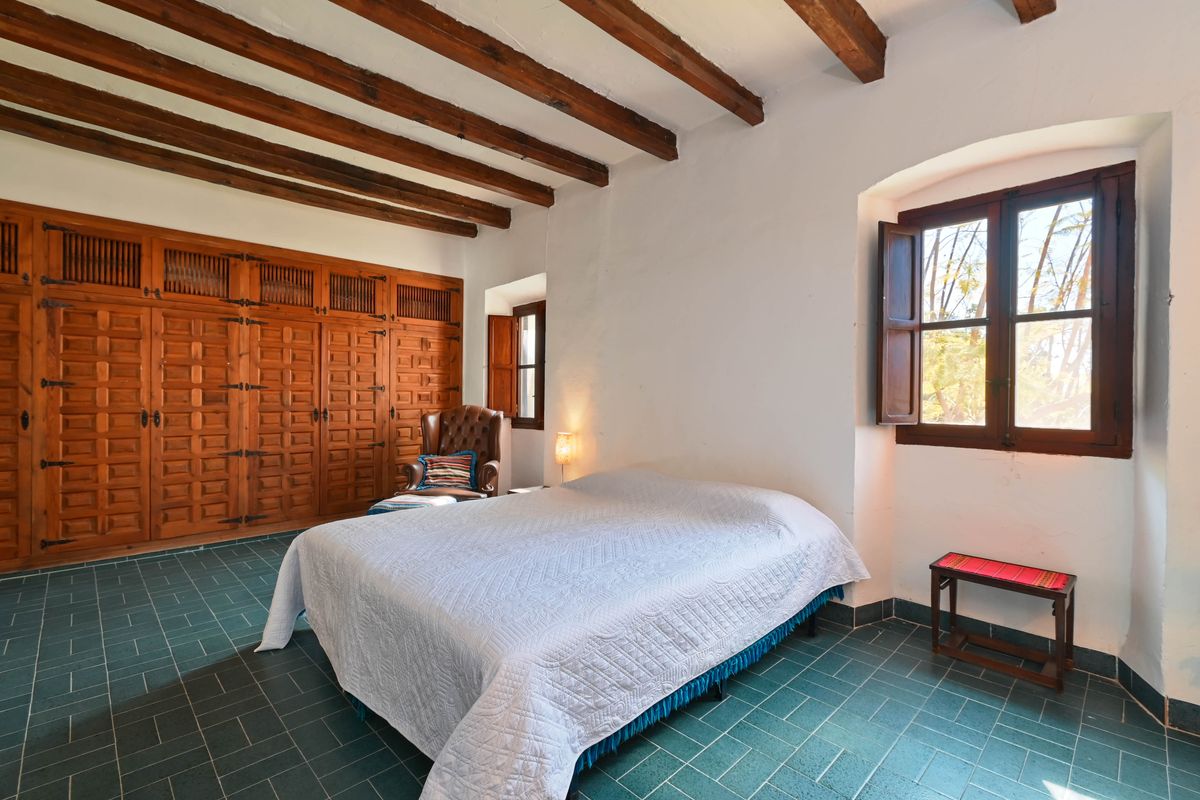 Dormitorio rústico con vigas de madera vistas, armarios empotrados de madera tallada, suelo de baldosas verdes y ventanas con contraventanas tradicionales.