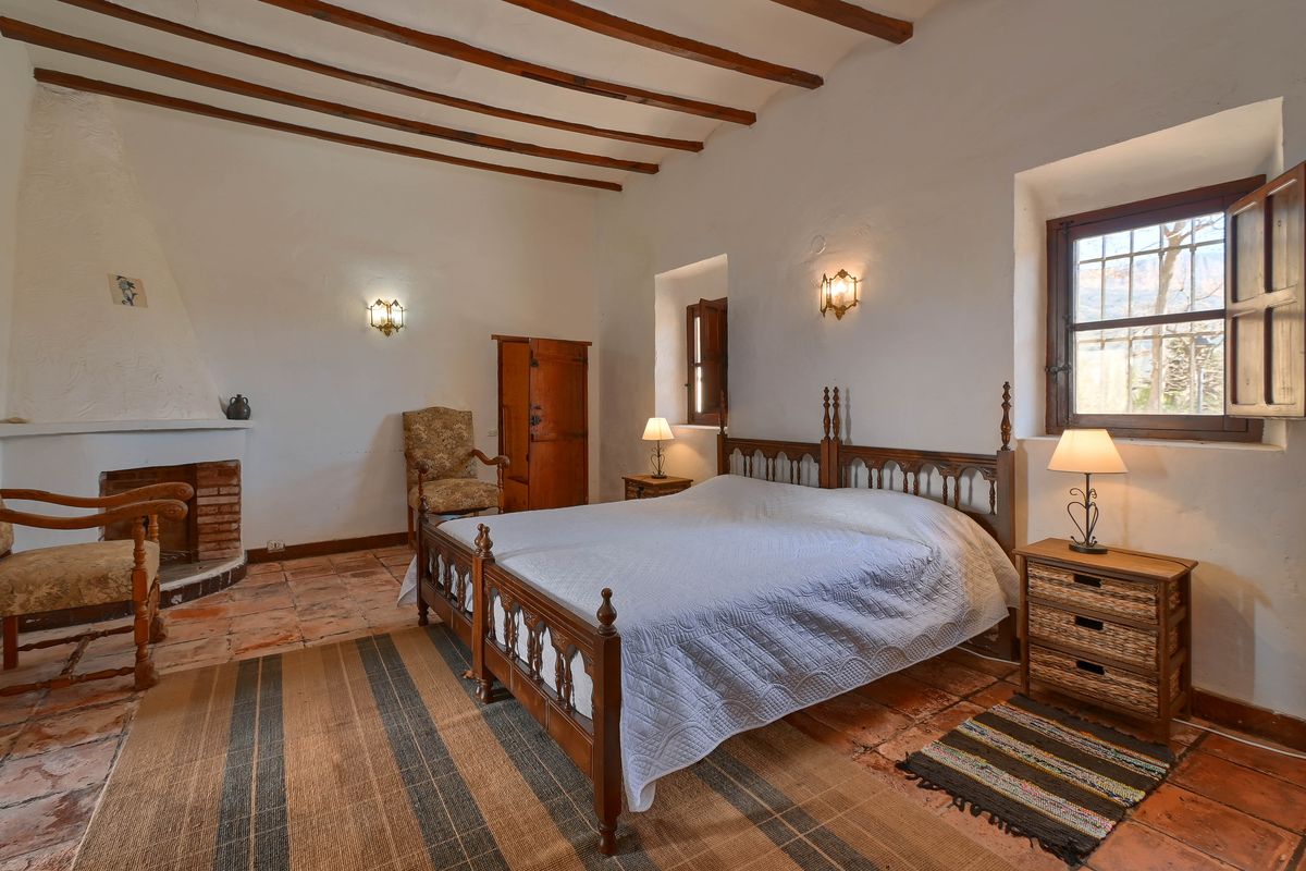 Dormitorio rústico con vigas de madera vistas, suelos de barro cocido, chimenea de obra y ventanas con contraventanas de madera tradicionales.