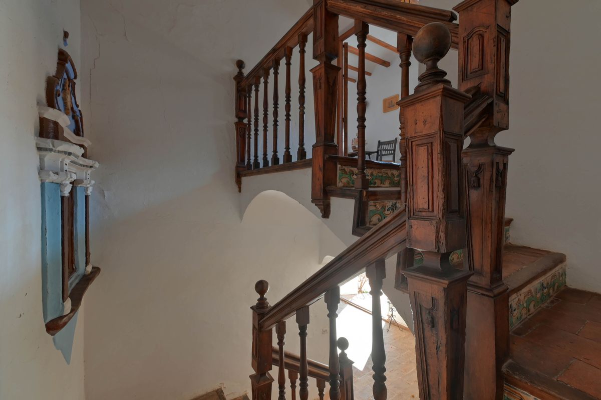 Escalera histórica de madera tallada con peldaños de barro cocido y contrahuellas de azulejos decorativos tradicionales sobre muros blancos.
