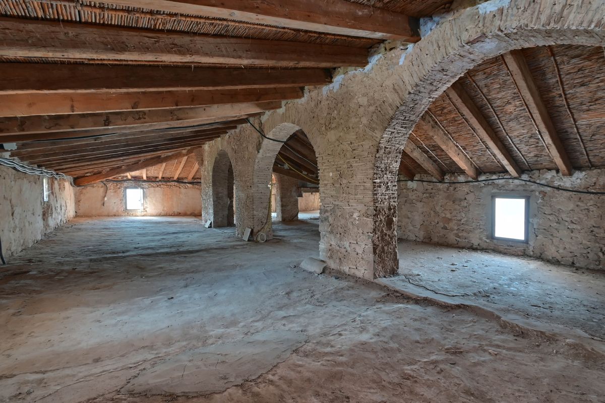 Espacio diáfano bajo cubierta con vigas de madera originales, techos de cañizo y arcos de piedra y ladrillo visto en estado original.