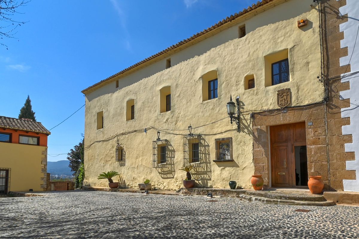 Fachada histórica con muros de textura ocre, portal de piedra tosca y escudo heráldico tallado. Destaca el patio de empedrado tradicional con macetas de barro cocido.