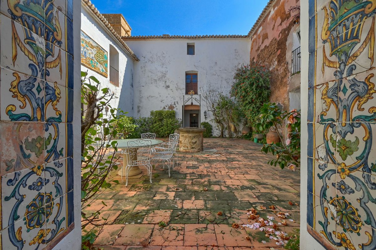 Patio interior histórico con suelos de terracota, pozo de piedra central y murales de azulejos decorativos en el arco de entrada.