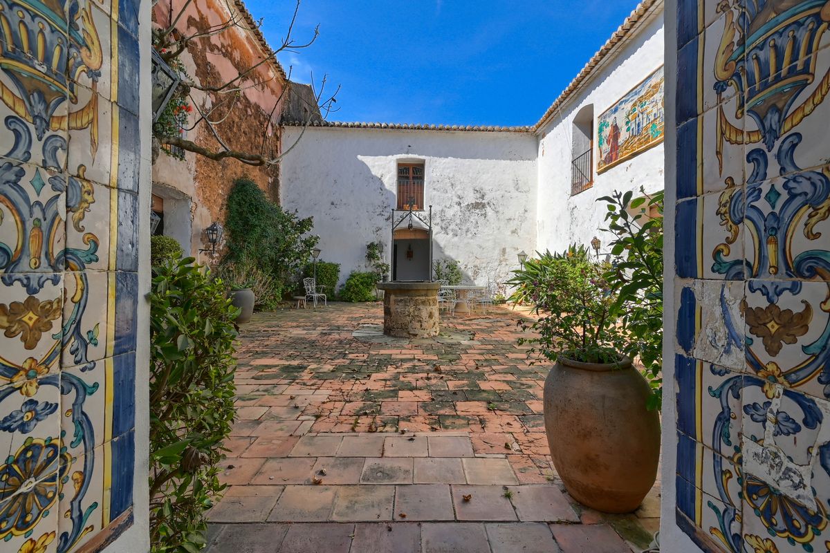 Patio interior tradicional con pozo central de piedra, suelos de barro cocido y azulejos decorativos pintados a mano en el arco de entrada.