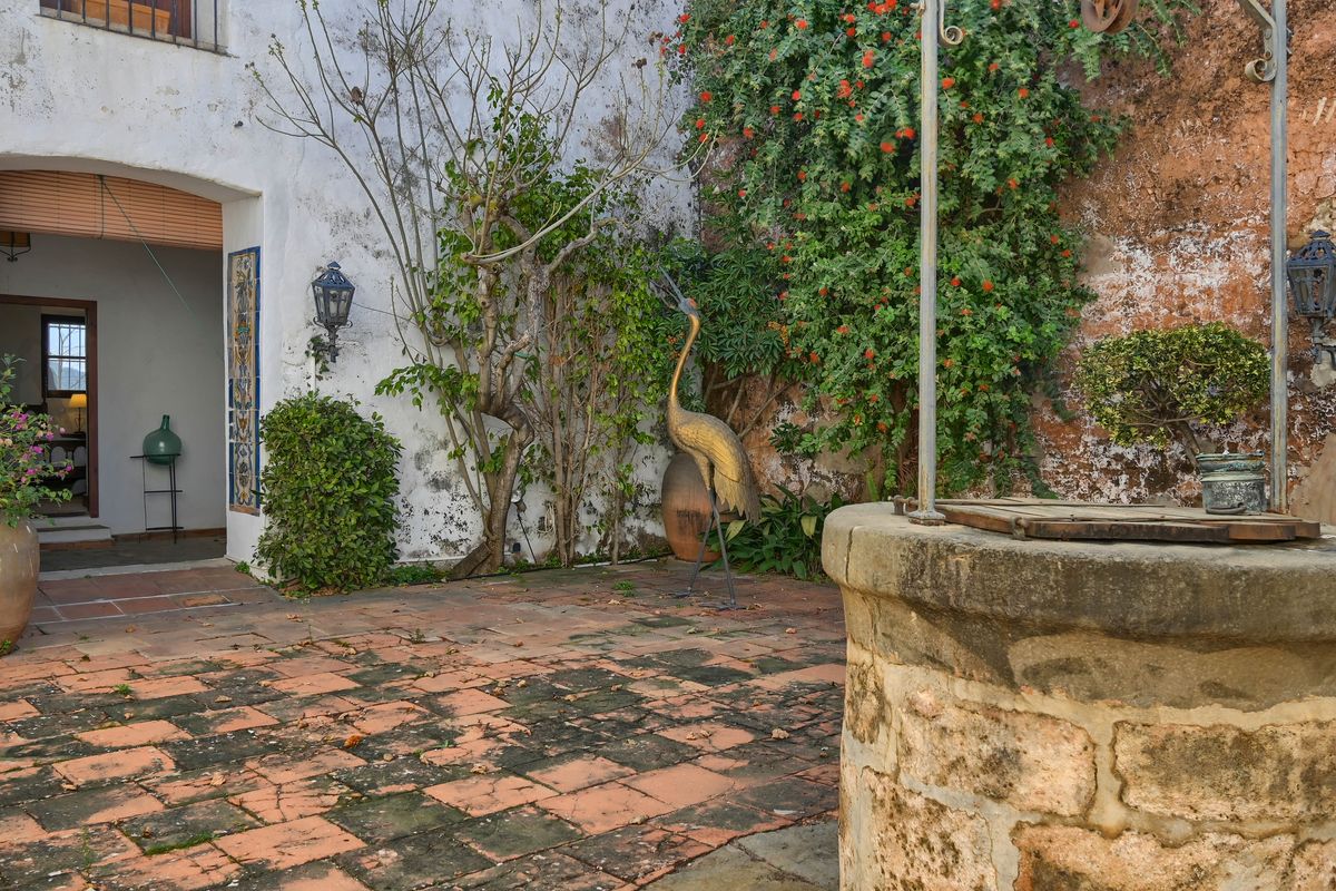 Patio mediterráneo tradicional con un pozo de piedra original, suelo de terracota envejecida y una escultura decorativa junto a muros encalados con vegetación.