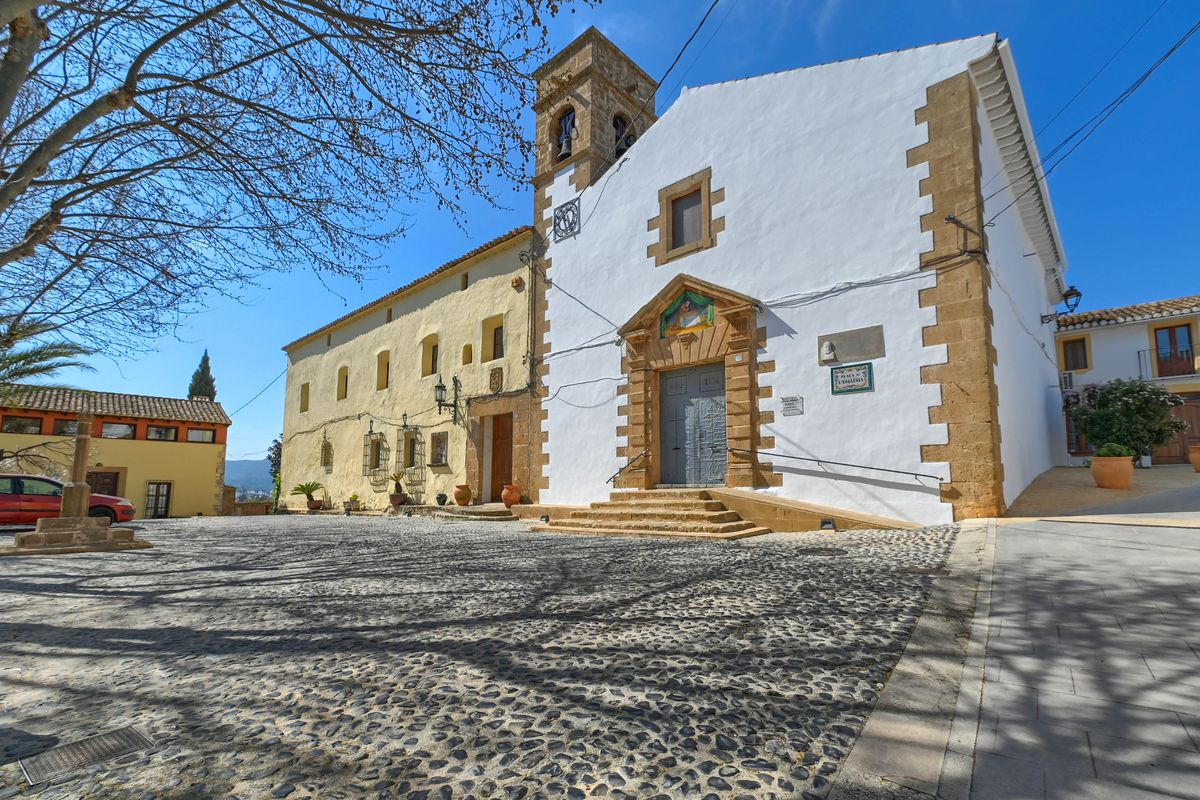 Plaza de la iglesia en Jesús Pobre con suelo de guijarros tradicionales, fachada blanca con sillares de piedra y campanario histórico bajo un cielo despejado.