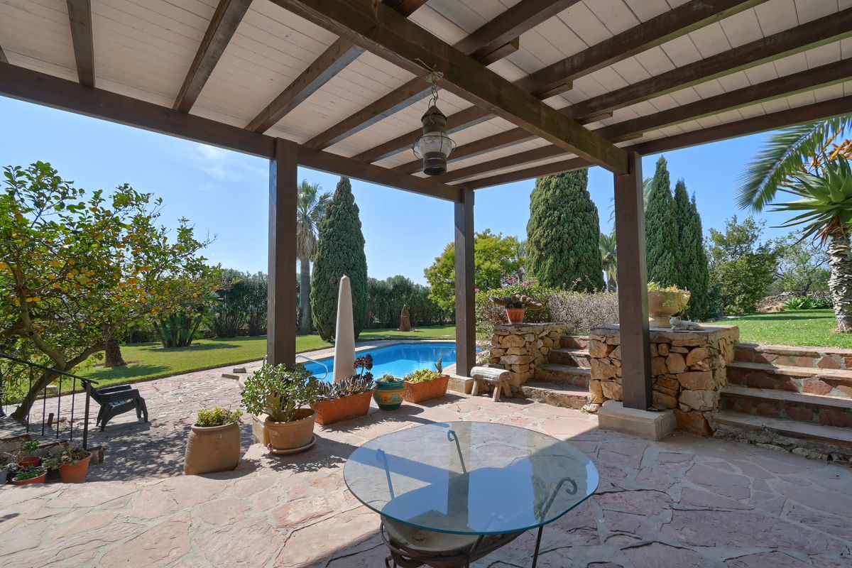 Porche cubierto con vigas de madera y suelo de piedra natural, con vistas directas a la piscina y al jardín mediterráneo.