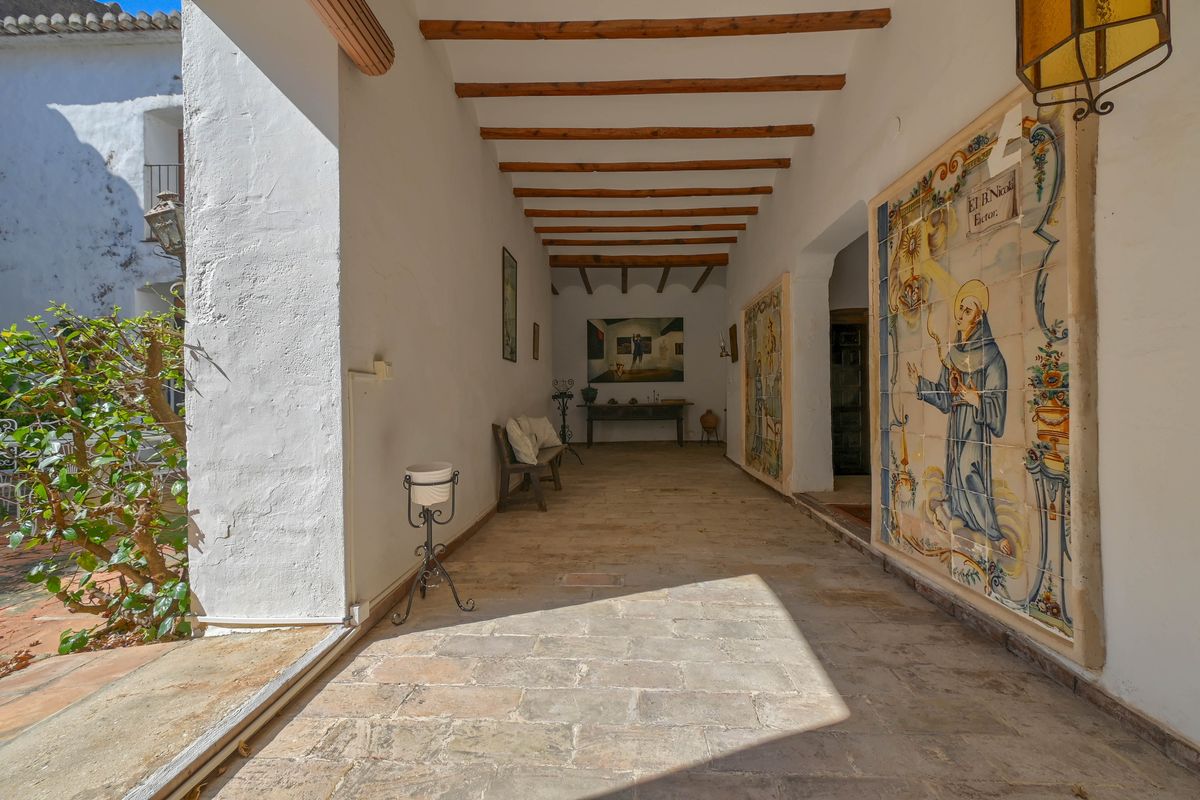 Porche cubierto tradicional con vigas de madera expuestas, suelos de piedra natural y un gran mural de azulejos religiosos históricos en la pared lateral.