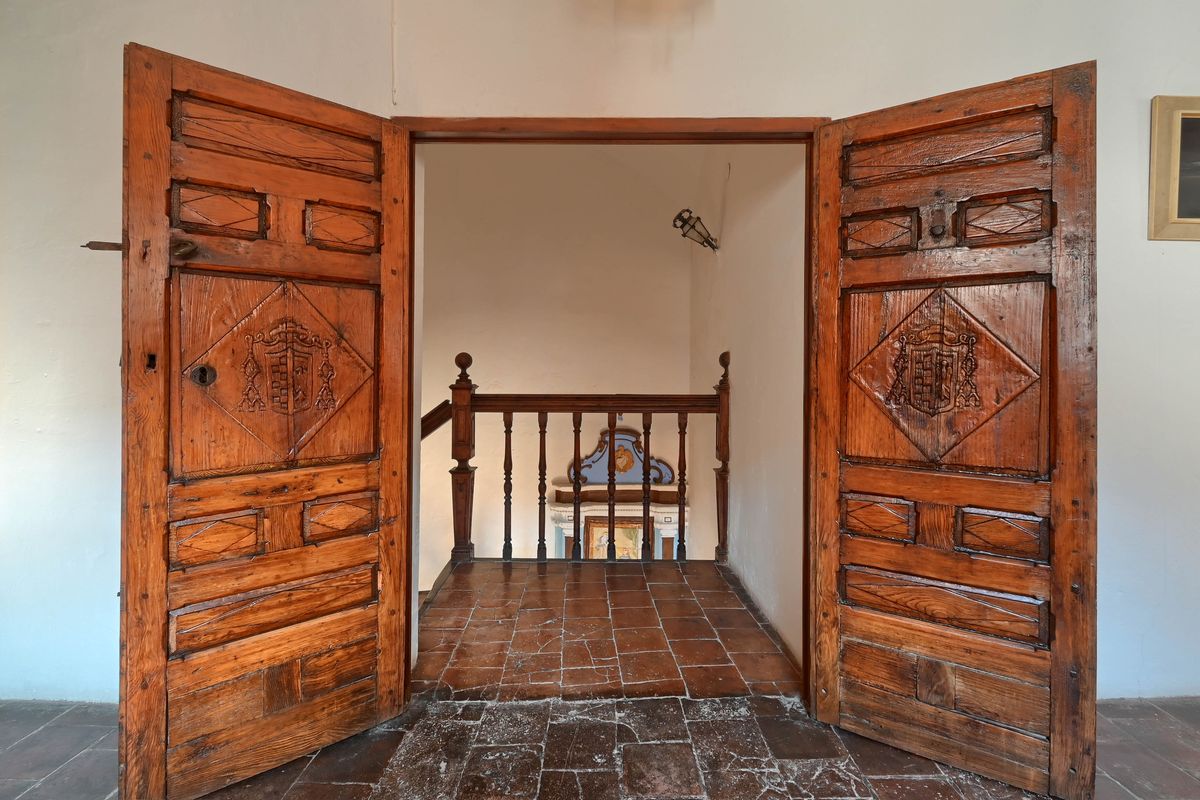 Puertas dobles de madera antigua con escudos heráldicos tallados que dan paso a un rellano con suelos de barro cocido y barandilla de madera.