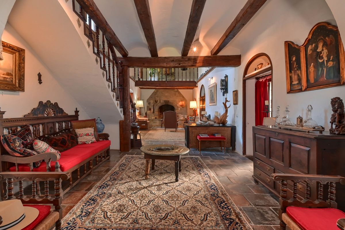 Recibidor tradicional con vigas de madera, suelos de barro cocido y altillo. Destacan los muebles de madera tallada y una chimenea de piedra al fondo.