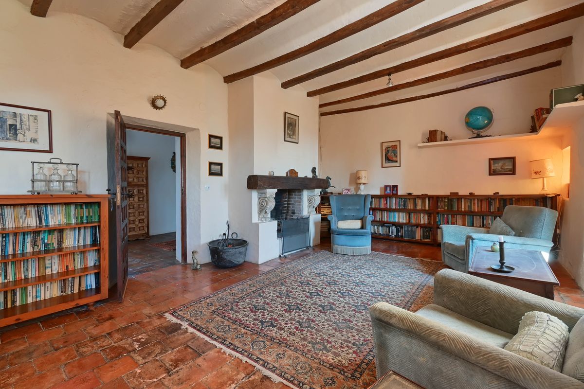Salón con vigas de madera vistas, chimenea de época con viga de madera, suelo de baldosa de barro cocido y estanterías de obra repletas de libros.