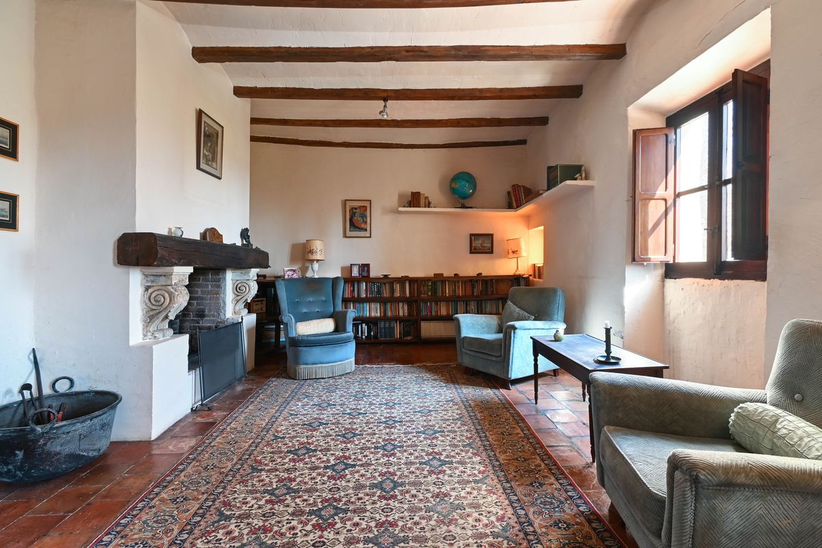 Salón histórico con vigas de madera vistas, chimenea con dintel de madera, suelos de barro cocido y estanterías integradas.