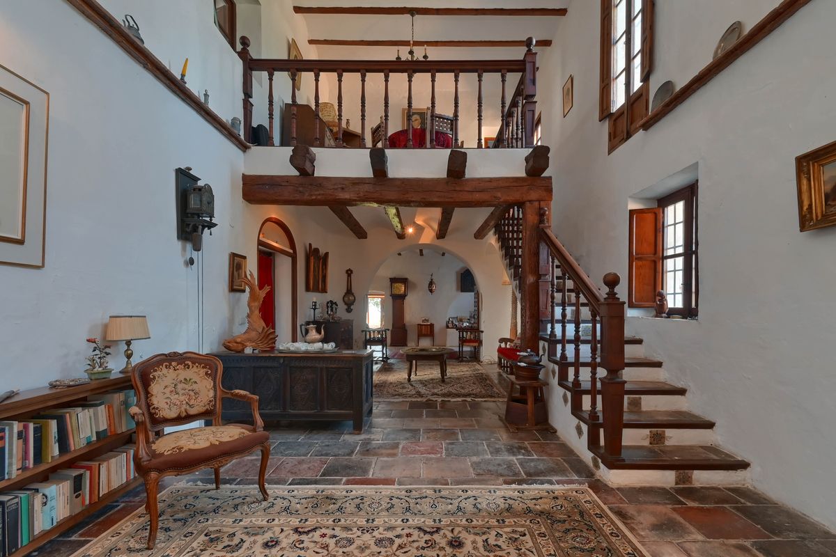 Salón principal de doble altura con vigas de madera vista, suelos de piedra rústica y una galería superior accesible por una escalera artesanal.