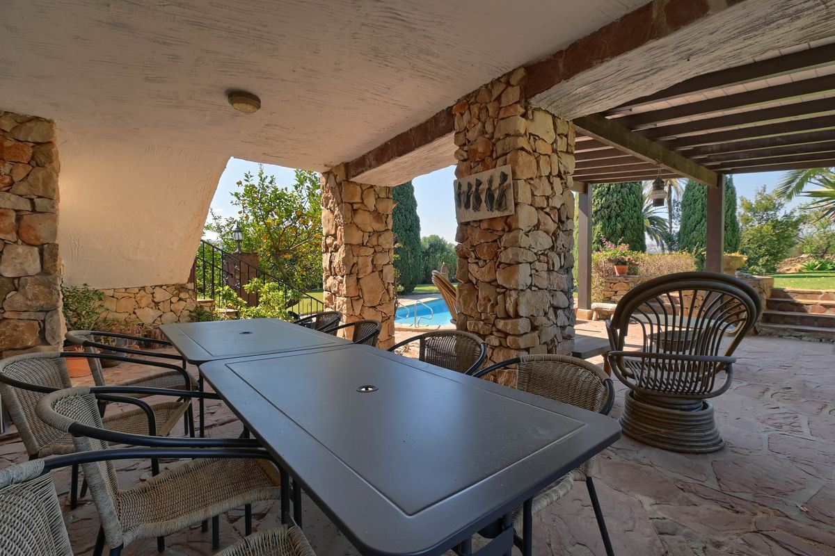 Terraza cubierta con pilares de piedra seca, comedor exterior y suelo de losa con vistas directas a la piscina y al jardín mediterráneo.