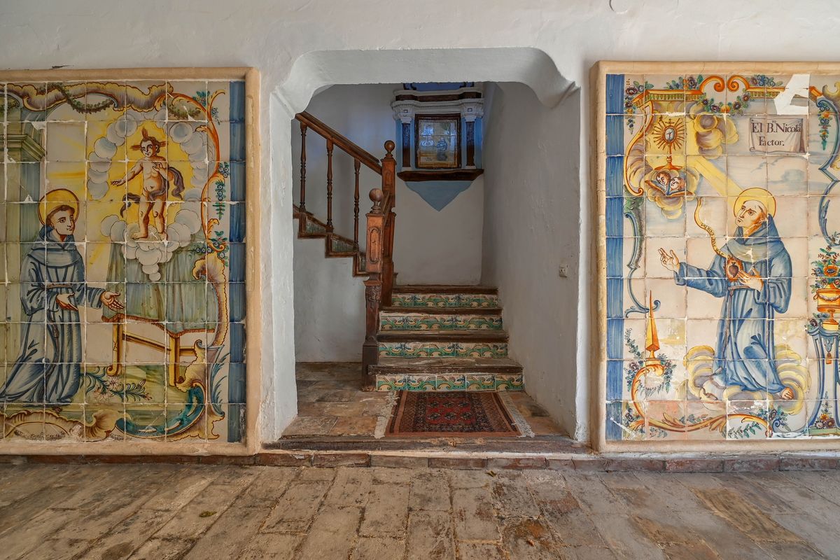 Zaguán con murales históricos de azulejos pintados a mano, muros de piedra encalados y escalera con frontales decorados y pasamanos de madera tallada.