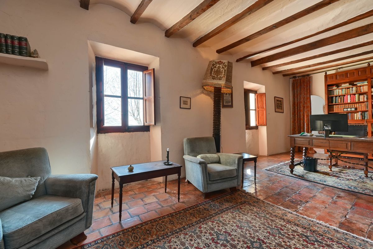 Zona de estudio con vigas de madera a la vista, suelos de terracota y ventanas profundas. Incluye un escritorio de madera antiguo y estantería integrada.