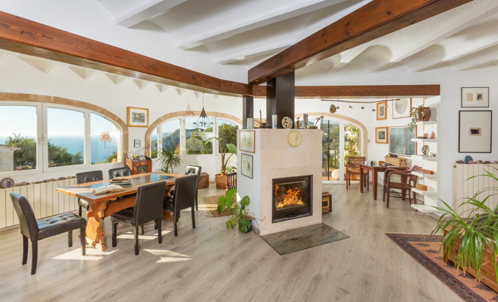 Amplio salón-comedor con chimenea central de piedra, vigas de madera oscura vista y ventanales en arco con vistas panorámicas al mar.