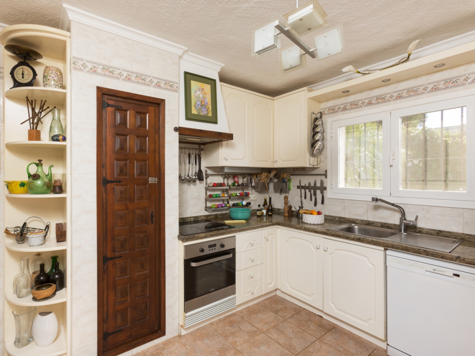 Cocina de estilo tradicional con armarios en tono crema, encimeras de granito y una puerta de despensa en madera oscura tallada. Incluye suelos cerámicos y ventana al exterior.