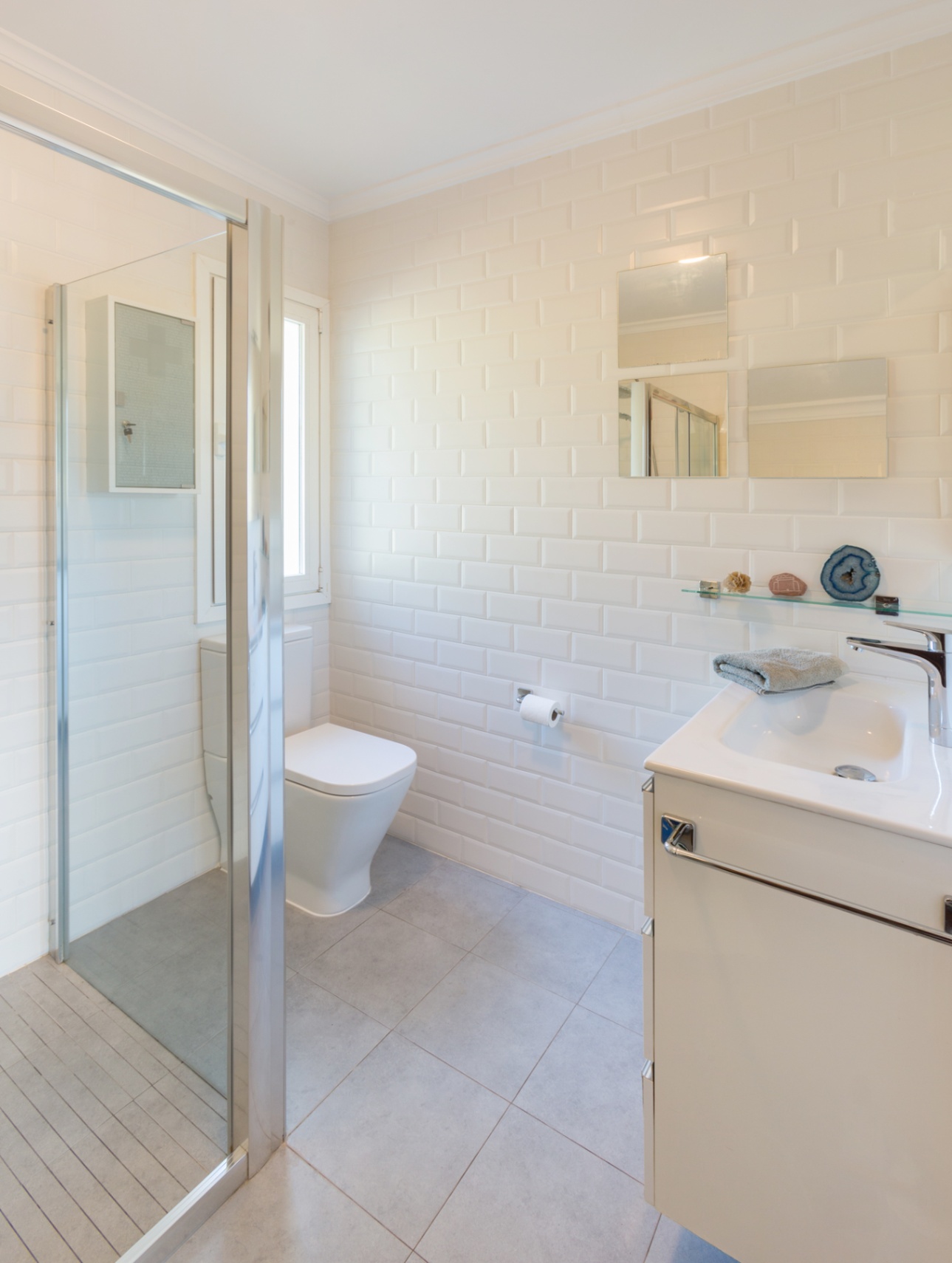 Cuarto de baño con azulejos tipo metro blancos, ducha con mampara de cristal y mueble lavabo minimalista. Incluye suelo cerámico gris y luz natural.