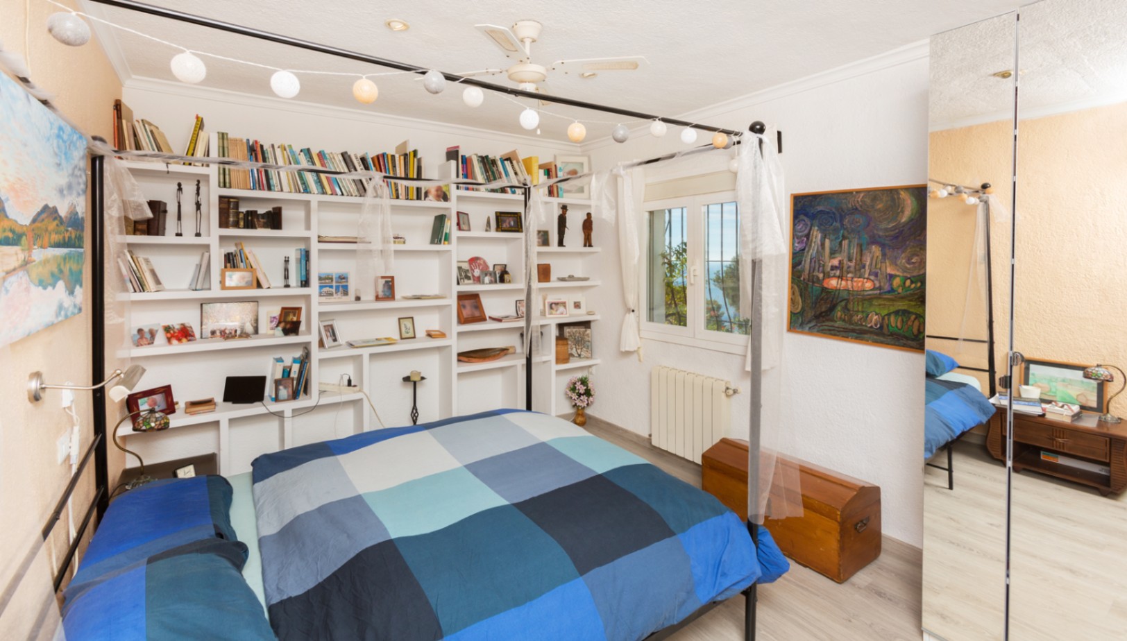 Dormitorio con cama de dosel de metal negro, estantería blanca integrada y armario con espejos. Dispone de ventilador de techo y ventana con vistas al Mediterráneo.