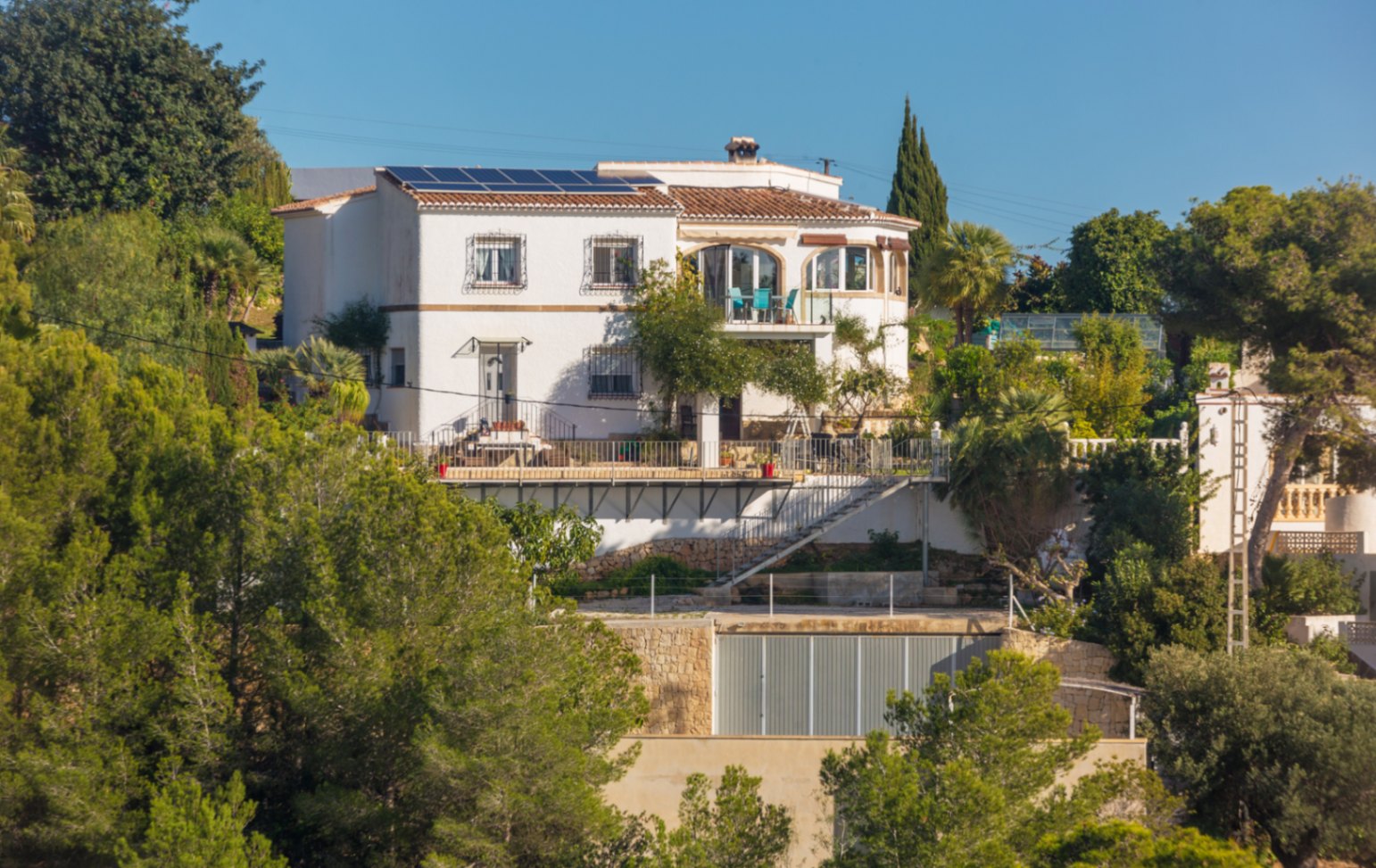 Fachada blanca de estilo mediterráneo con paneles solares, terrazas exteriores y muros de piedra, integrada en una ladera con abundante vegetación local.