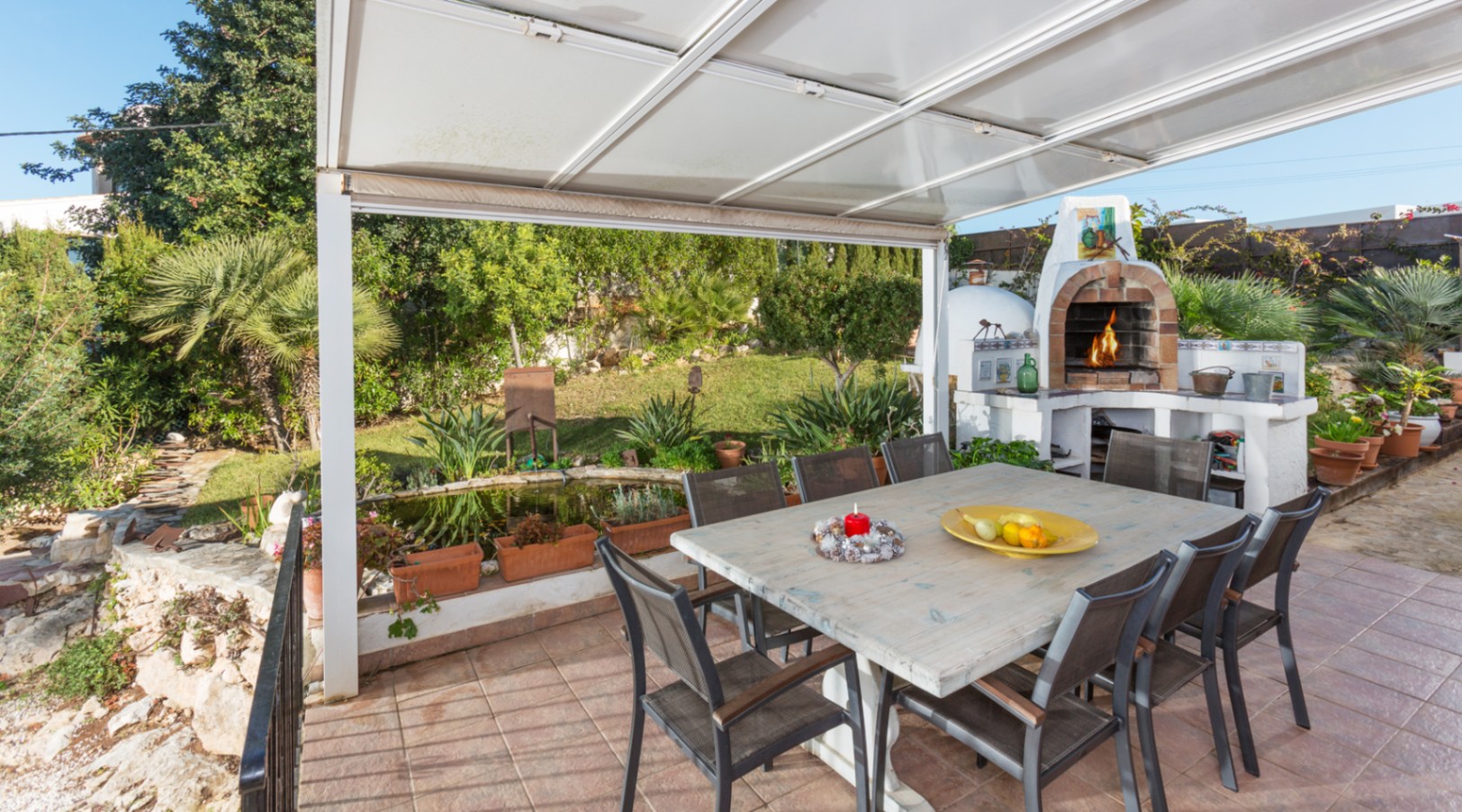 Terraza cubierta con amplia mesa de madera, horno de leña tradicional de ladrillo y vistas al jardín mediterráneo con estanque privado.