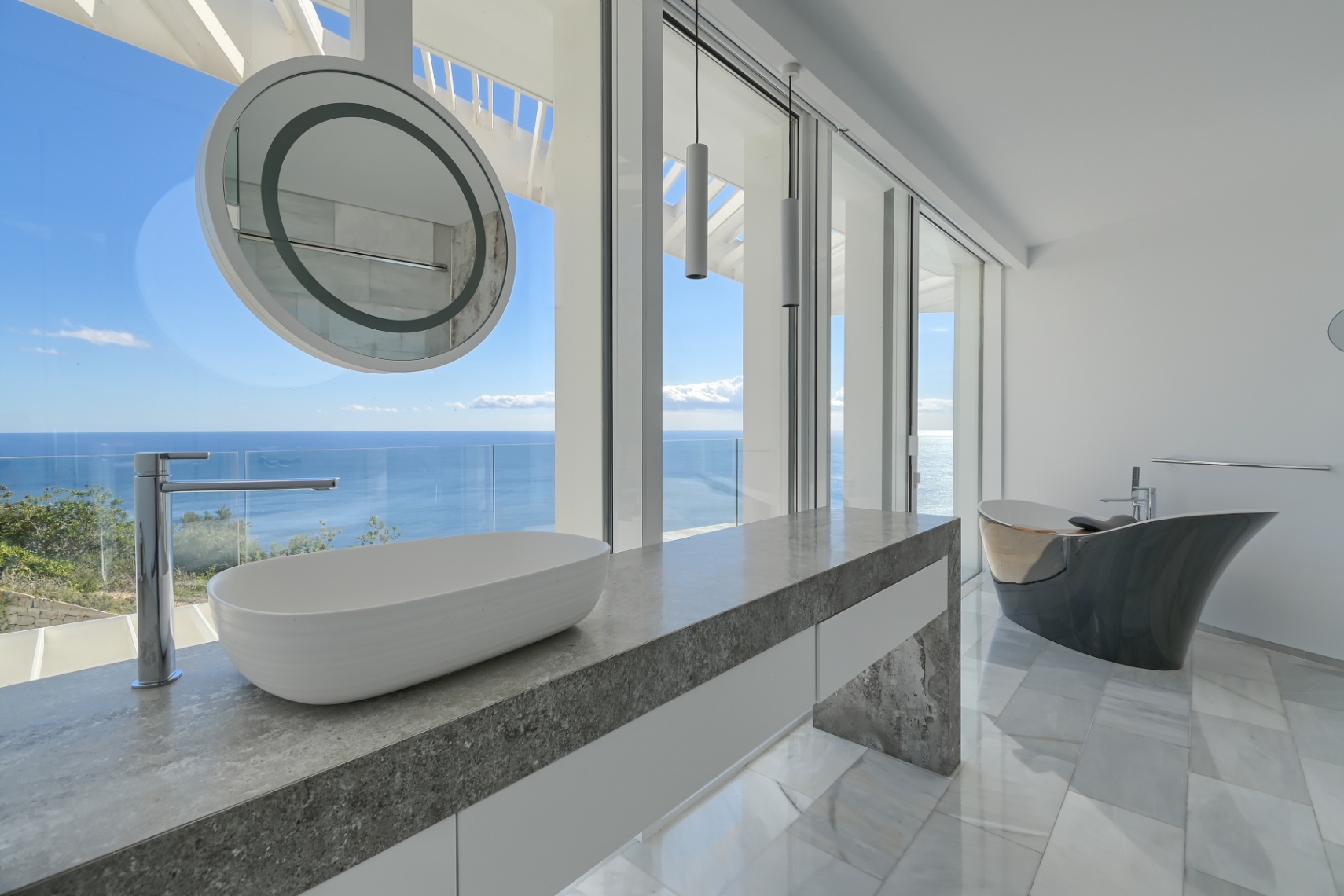 Baño moderno con encimera de piedra gris, lavabo sobre encimera y bañera exenta de diseño, con ventanales de suelo a techo y vistas panorámicas al mar.