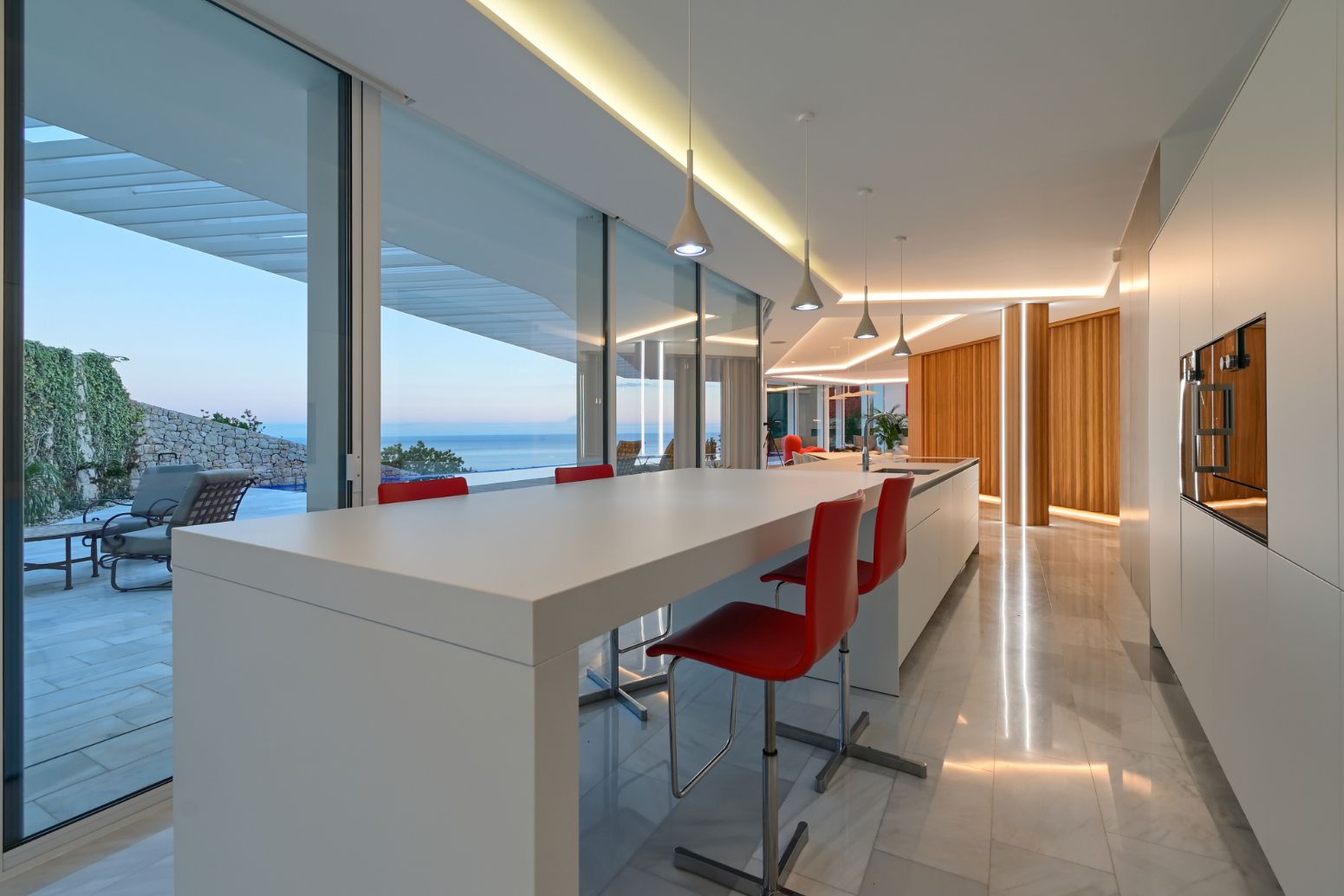 Cocina moderna con isla blanca minimalista y taburetes rojos, ventanales de suelo a techo con vistas al mar e iluminación LED integrada.