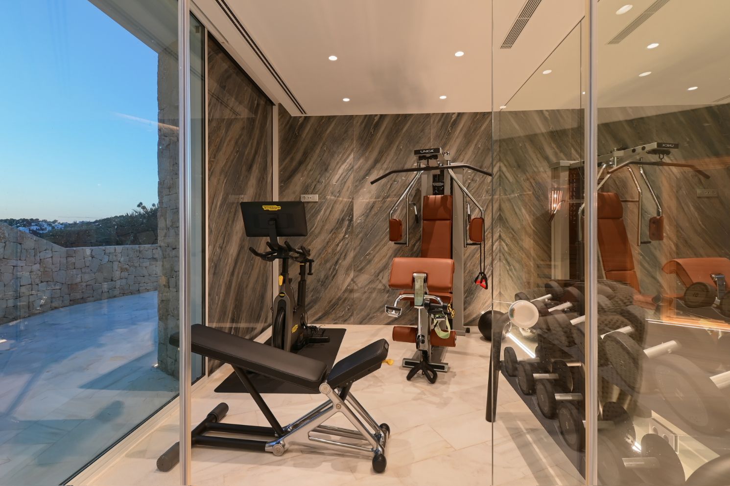 Gimnasio privado con equipamiento Technogym, paredes revestidas en piedra veteada y amplios ventanales con vistas a una terraza de piedra natural.