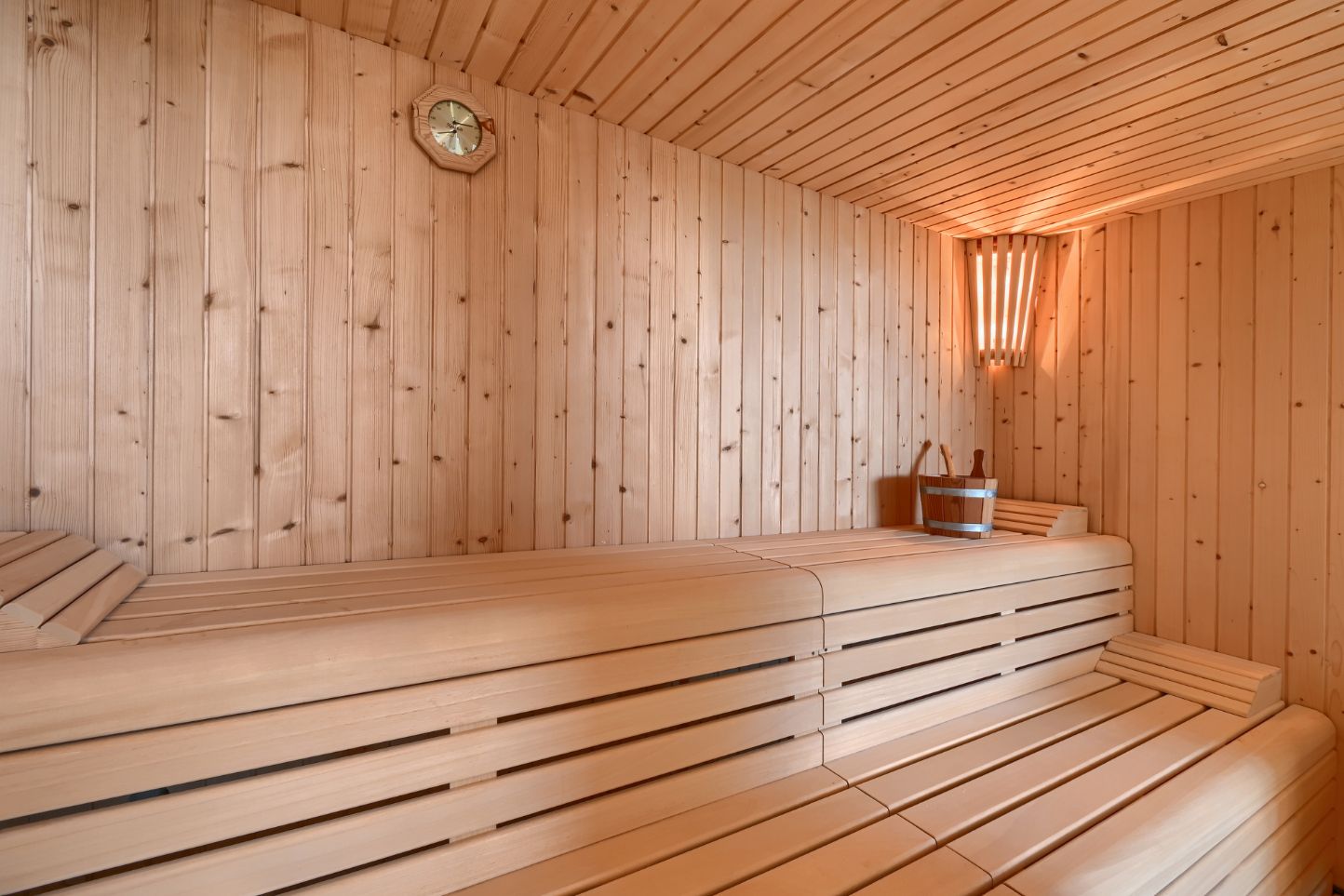 Interior de sauna con revestimiento de madera clara, bancos de listones a varios niveles, aplique de luz en esquina y accesorios de madera tradicionales.
