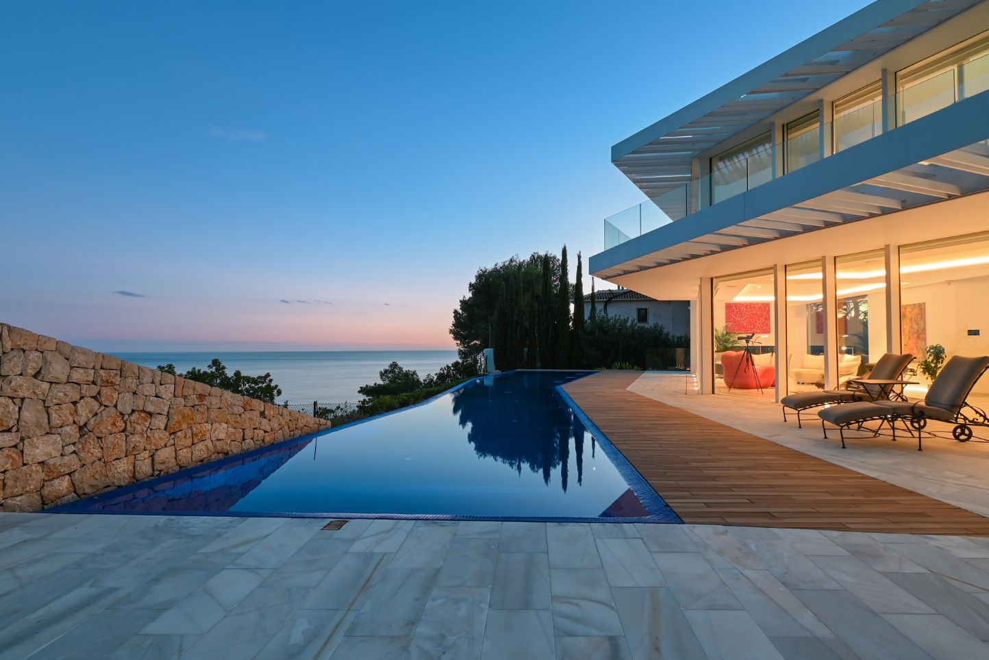 Piscina infinita con tarima de madera y pavimentos de piedra frente al mar. La villa moderna destaca por sus amplios ventanales y muros de piedra natural.