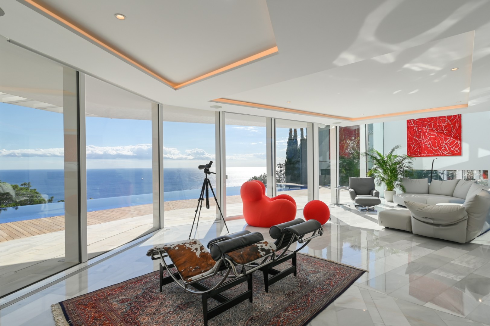Salón luminoso con acristalamiento de suelo a techo, suelos de mármol y asientos de diseño con vistas panorámicas a la piscina infinita y al mar.