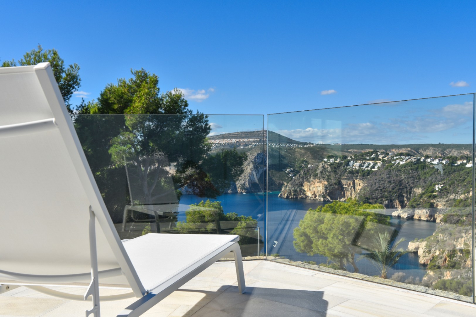Terraza con tumbona blanca y barandilla de cristal sin marcos, con vistas panorámicas a una bahía mediterránea y acantilados con pinos.
