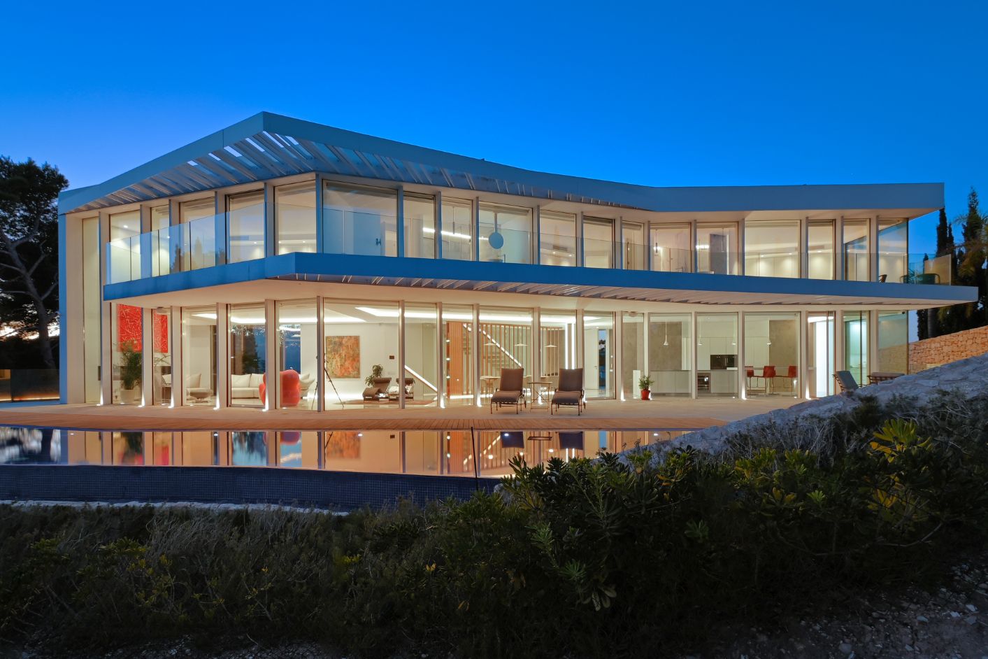 Villa moderna de dos niveles con fachadas acristaladas, amplia terraza con piscina reflectante e iluminación interior integrada al anochecer.