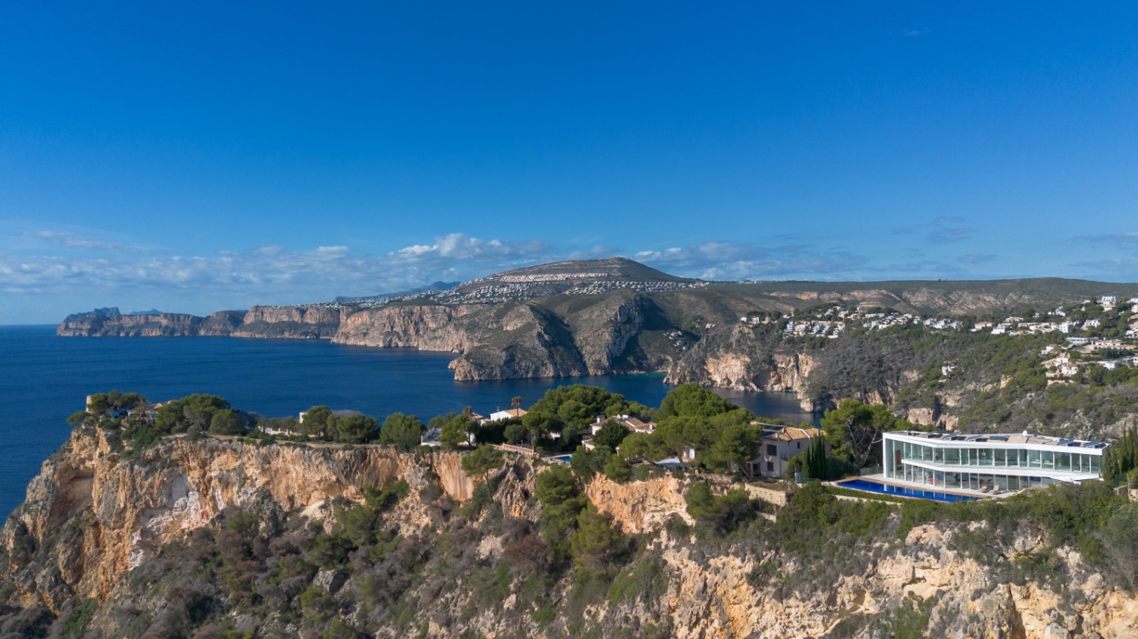 Vista aérea de una villa contemporánea con fachada de vidrio en un acantilado, destacando su piscina lineal y vistas despejadas al mar Mediterráneo.