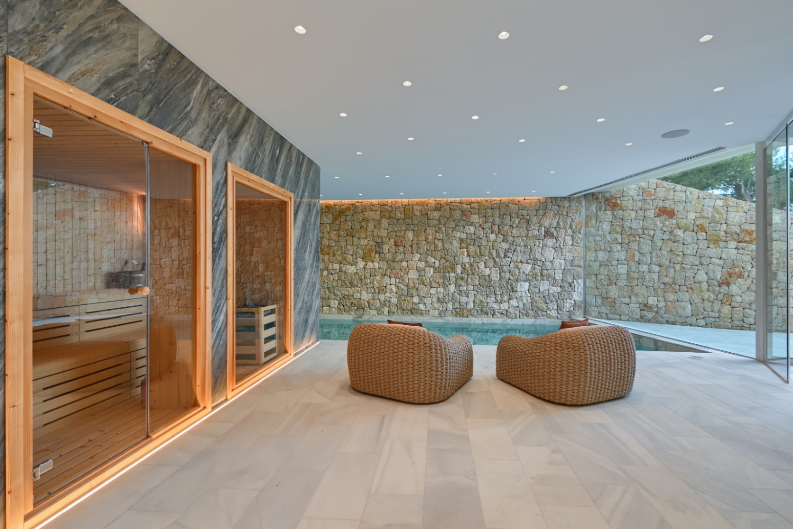 Zona de spa interior con sauna de madera, sillones de fibras naturales y paredes de piedra rústica frente a una piscina privada integrada.