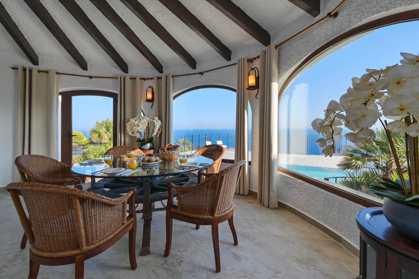 Comedor circular con mesa de cristal sobre base de rueda de carro y sillas de mimbre, bajo un techo de vigas vistas con amplios ventanales arqueados hacia el mar.