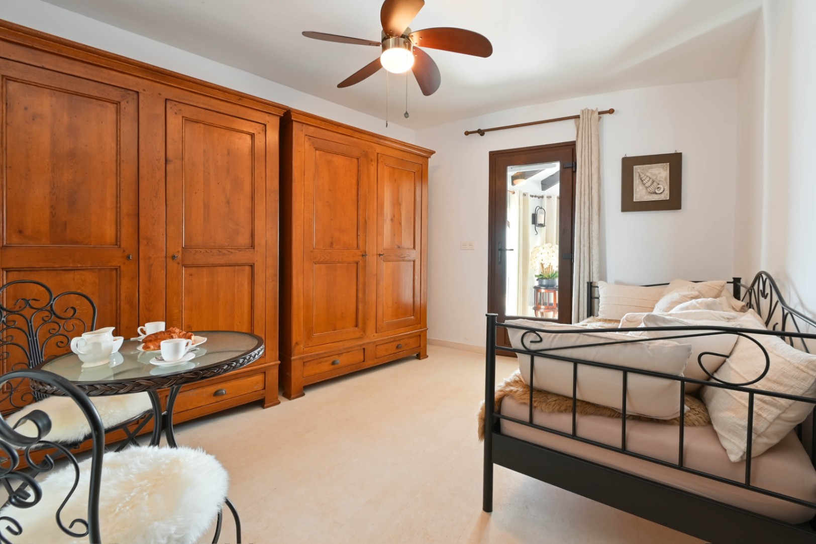 Dormitorio con dos grandes armarios de madera rústica, cama de forja negra y zona de estar tipo bistró. Dispone de ventilador de techo y puerta acristalada al exterior.