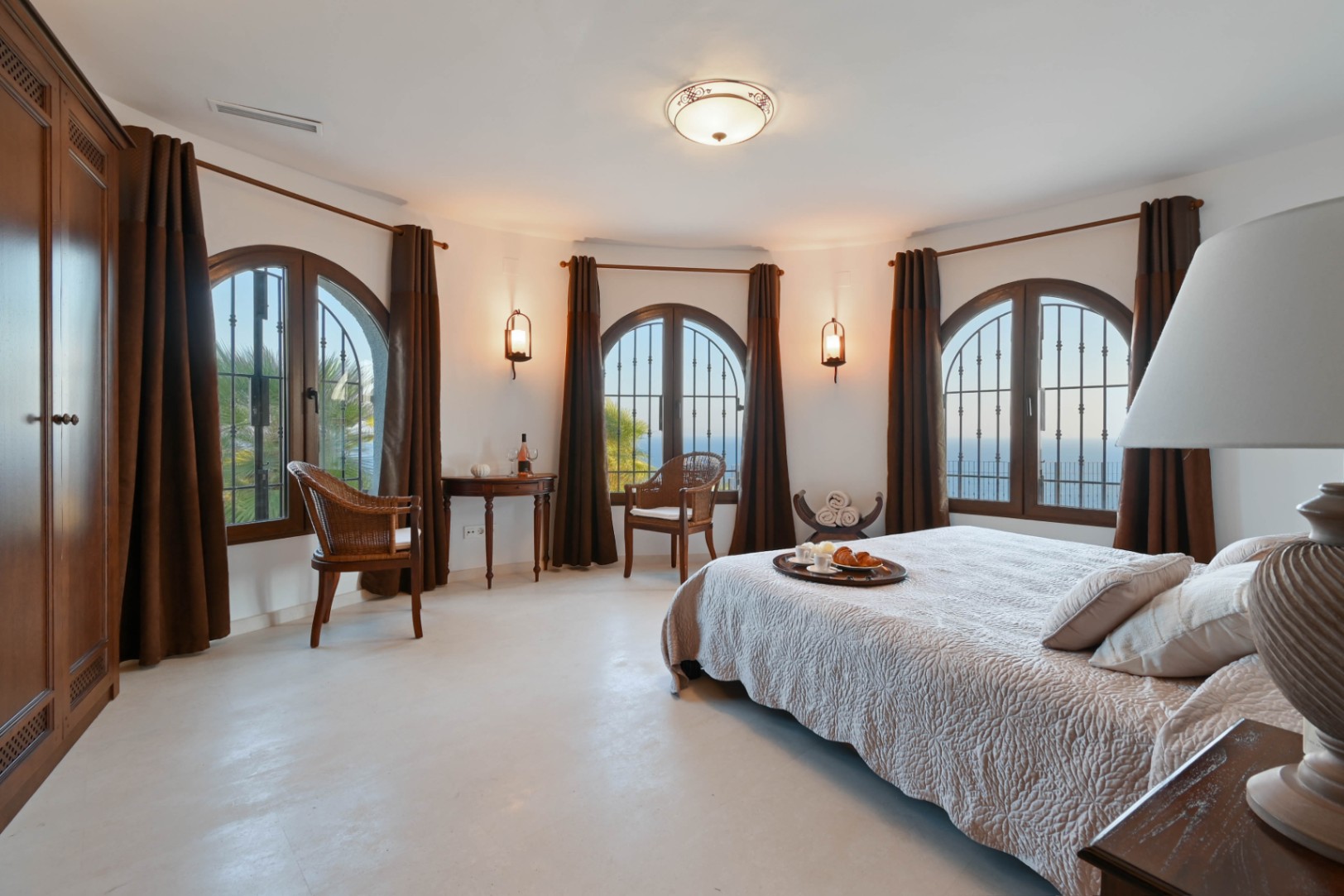 Dormitorio principal con pared curva, tres ventanales de medio punto en madera oscura, vistas al mar y mobiliario de mimbre y madera.