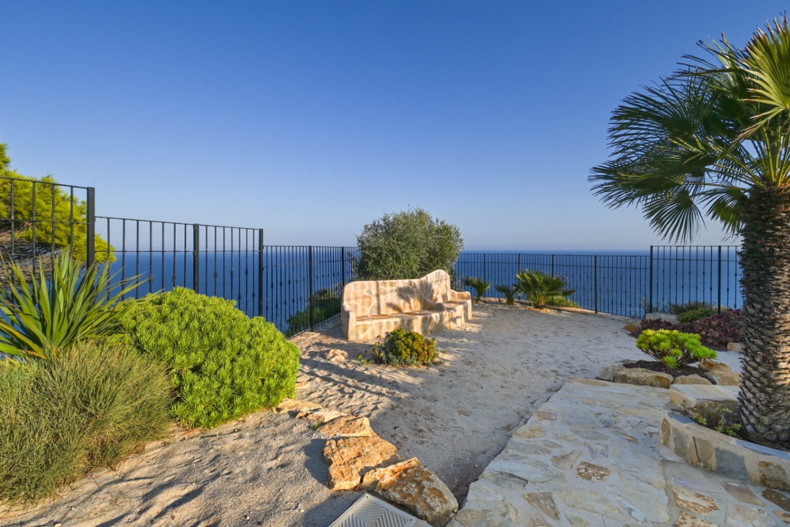 Mirador exterior con banco de piedra natural, vallado de forja negra y vistas despejadas al mar Mediterráneo rodeado de vegetación autóctona.